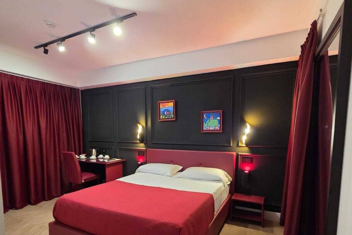 A modern hotel room with a large bed, red curtains, a desk with a chair, and two wall-mounted lamps.