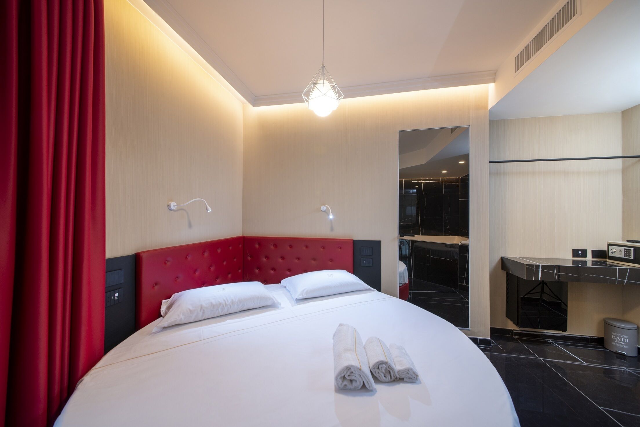 Une chambre d’hôtel avec un grand lit, des rideaux rouges, une tête de lit moderne, une table de chevet et un miroir.