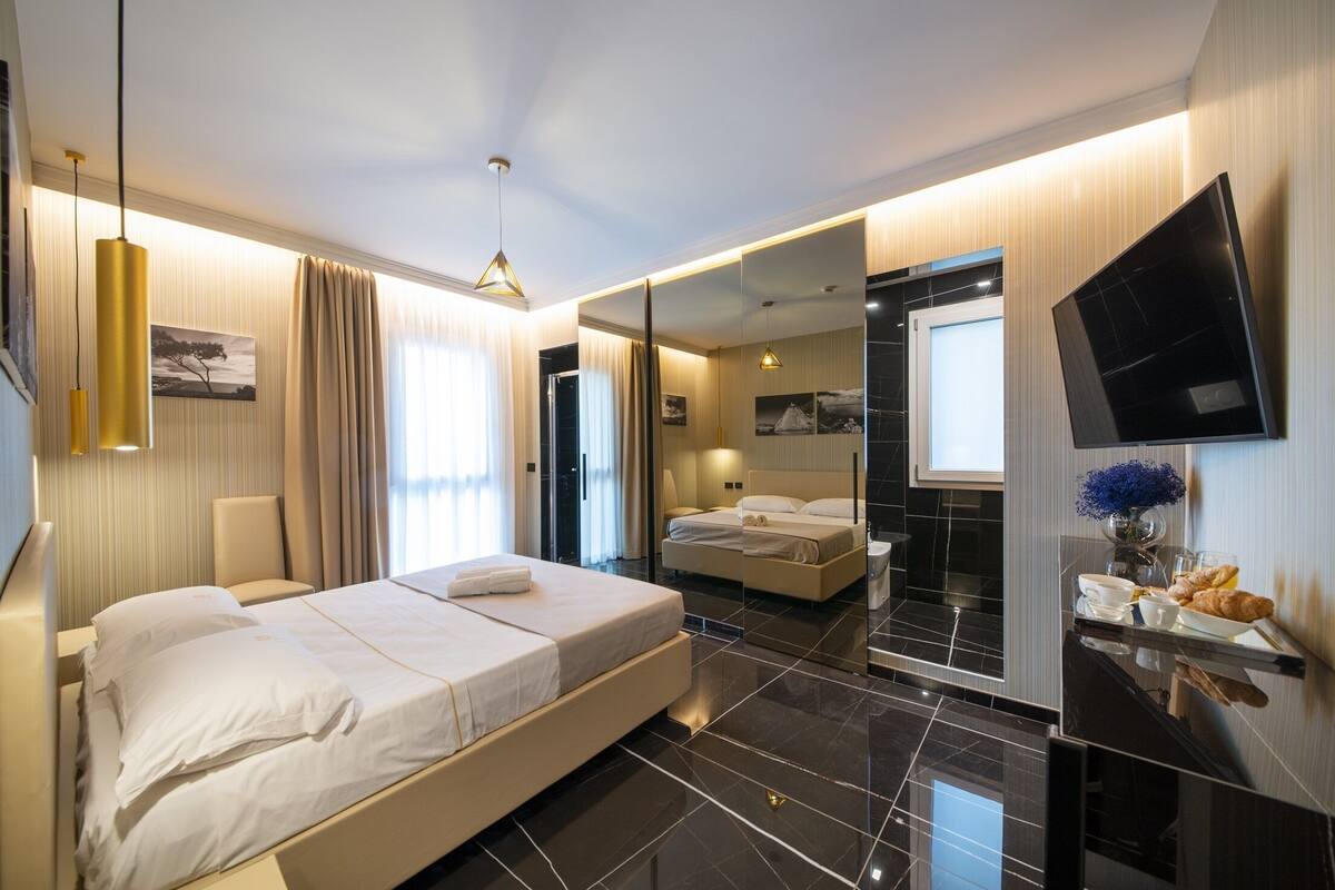 A modern hotel room with a large bed, a TV, a mirror, and a dining area with a vase of flowers.