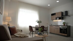A modern living room with a sofa, coffee table, and a dining area with chairs. There is a kitchen with wooden cabinets, a microwave, and a stove. - X Business (Saviano)