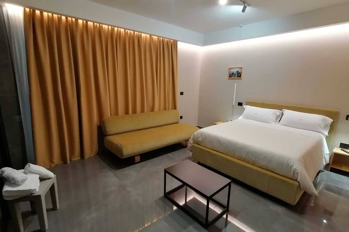 A modern hotel room with a large bed, a sofa, a small table, and a curtain.