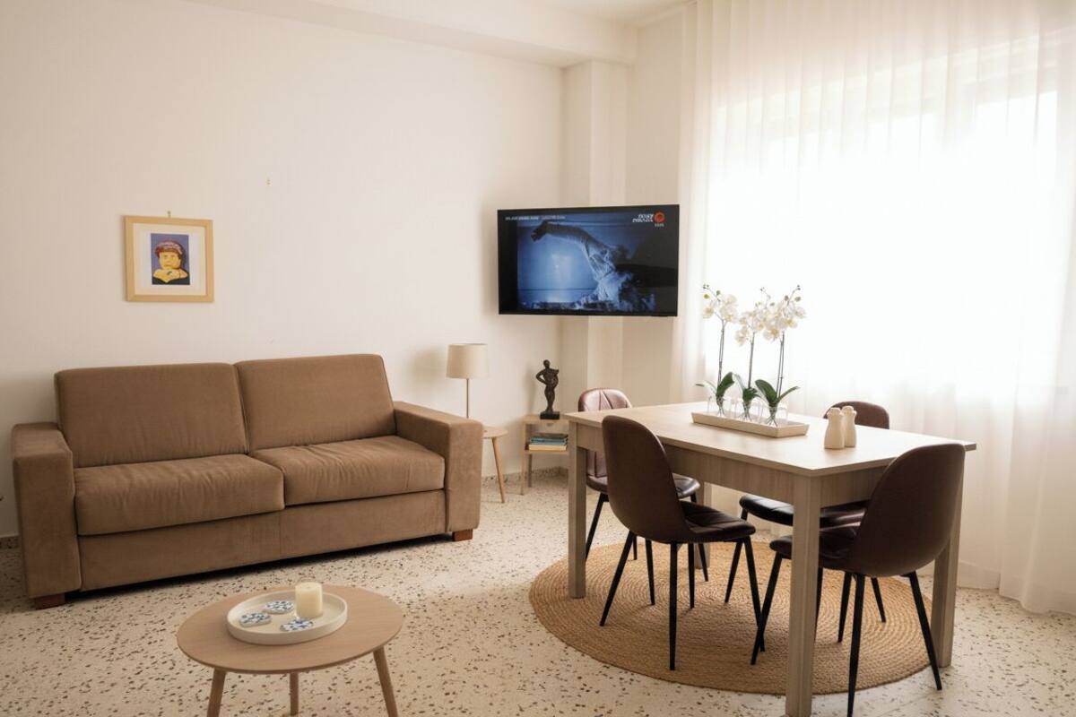 A living room with a beige sofa, a coffee table, a TV mounted on the wall, and a dining area with a table and chairs.