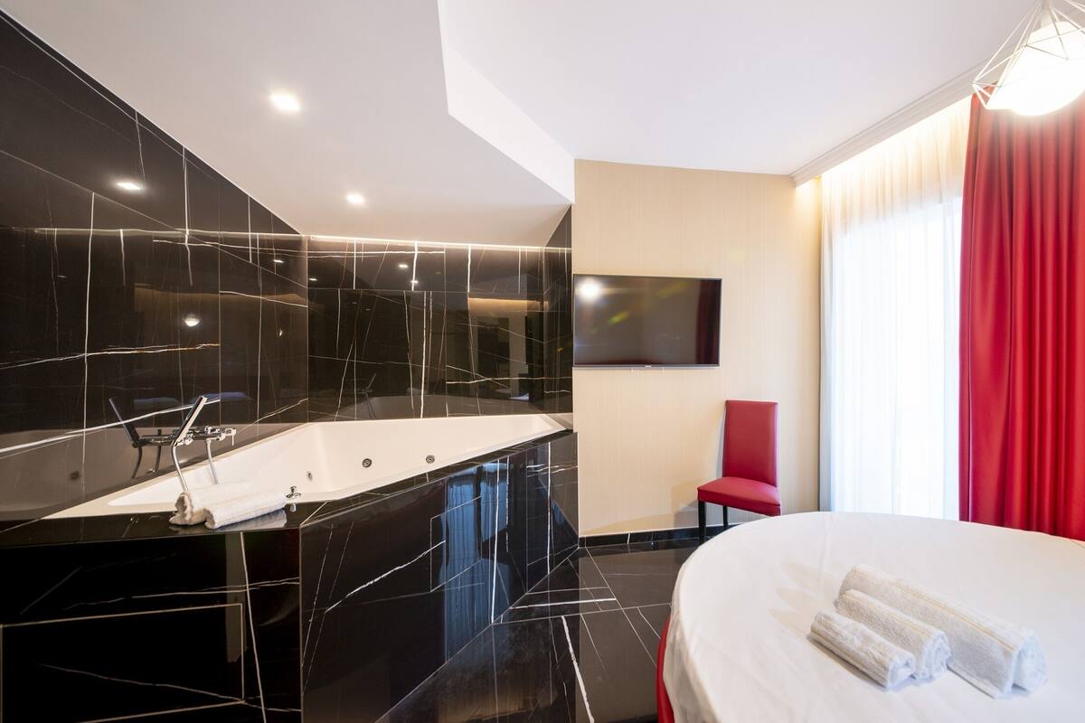 A modern hotel room with a large black marble bathtub, a white bed, and a red chair.