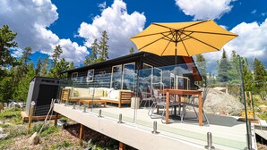 A modern log cabin with a glass enclosure, outdoor seating, and a large yellow umbrella. - BeeHive Bungalow - Gorgeous Lake views and completely updated throughout! (Grand Lake)