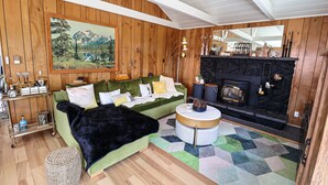 A cozy living room with a green sofa, a fireplace, a painting of a mountain landscape, and a wooden ceiling. - BeeHive Bungalow - Gorgeous Lake views and completely updated throughout! (Grand Lake)