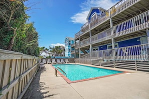 A pool area with a wooden fence, lounge chairs, and a multi-story building with balconies.