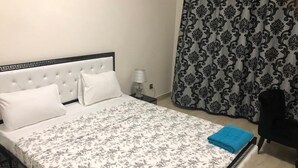 Ein ordentlich bezogenes Bett mit floralem Überwurf, eine Nachttischlampe und ein schwarzer Stuhl in einem Raum mit gemusterten Vorhängen.