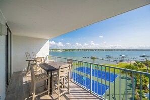 A balcony with a table and chairs, overlooking a tennis court and a body of water with docks. - Gulf Shores Surf and Racquet Club 502B - 2 Bed Condo (Gulf Shores)