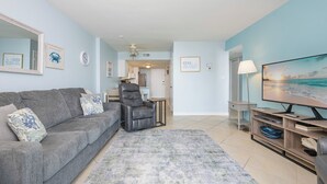 Een woonkamer met een grijze bank, een zwarte fauteuil, een televisie en een schilderij van een strandgezicht.