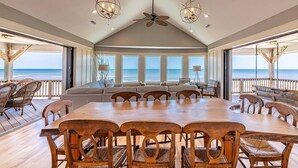 Een ruime eetruimte met een grote houten tafel en stoelen, een plafondventilator en grote ramen met uitzicht op de oceaan.