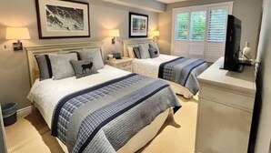 A hotel room with two beds, a desk, a TV, and framed pictures on the wall.