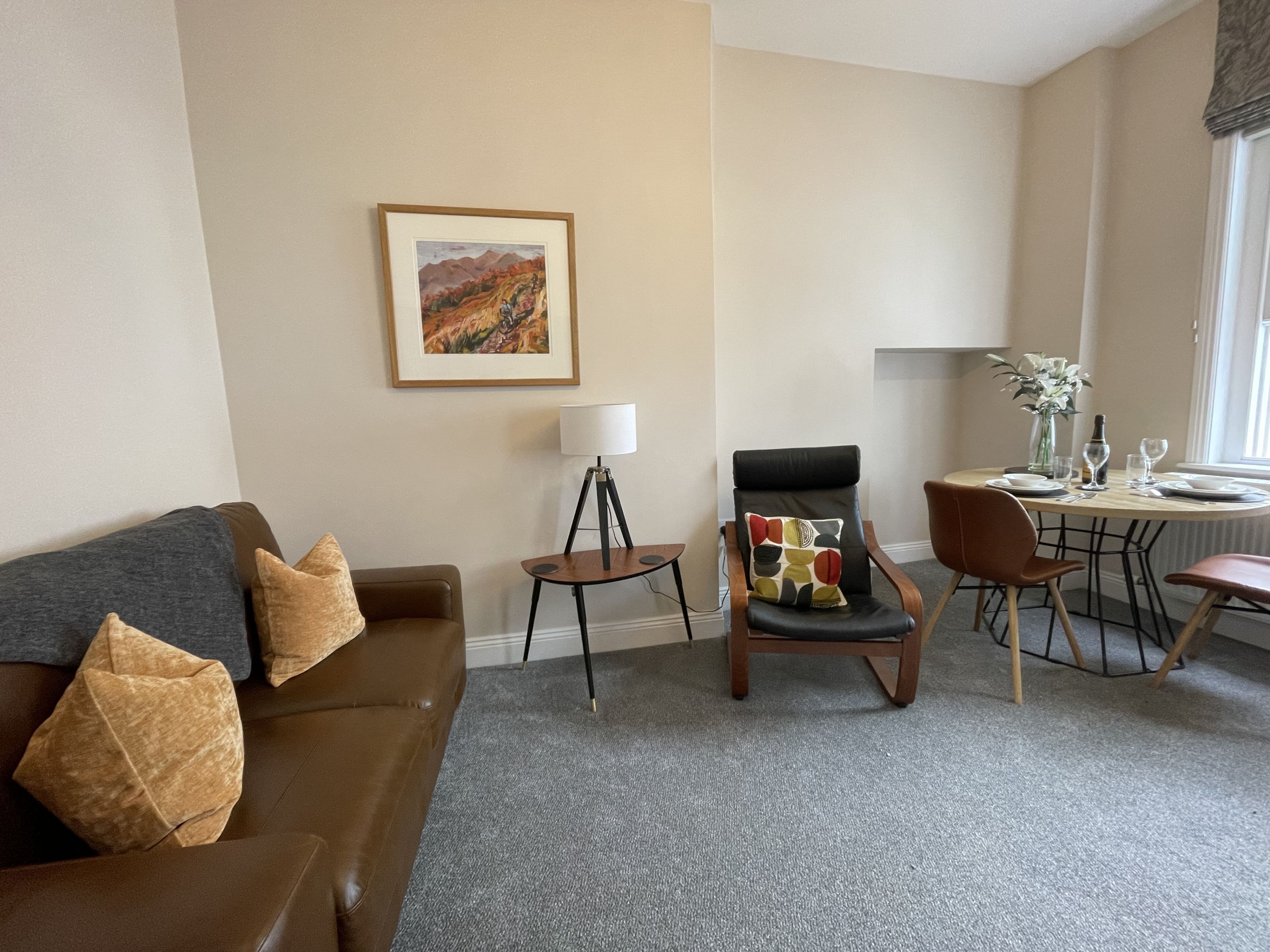Quarto de hotel com sofá de couro marrom, mesinha lateral com abajur, mesa de jantar posta para dois e uma pintura emoldurada de paisagem na parede.