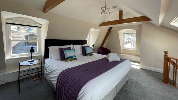 A bedroom with a large bed, a small table, a chair, and a window with a view of buildings. - Grisedale Pike -  a maisonette that sleeps 2 guests  in 1 bedroom (Central Keswick)