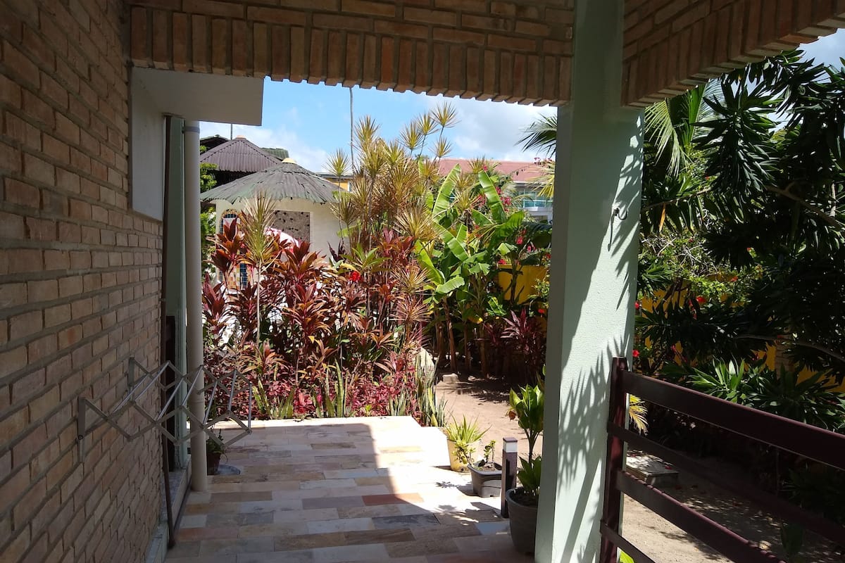 A covered outdoor area with a tiled floor leading to a pathway surrounded by lush greenery and potted plants.