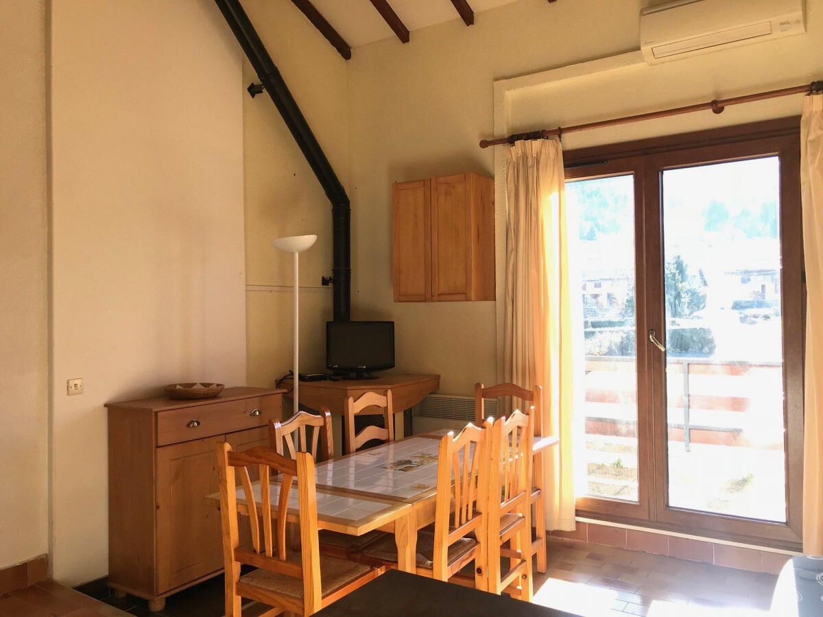Una habitación acogedora con una mesa de comedor de madera, sillas, un armario, televisión y un ventanal con cortinas.