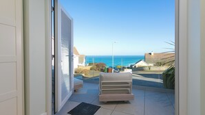 Ein Balkon mit einer weißen Couch, einer schwarzen Fußmatte und freiem Blick auf das Meer und Gebäude in der Ferne.