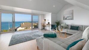Ein geräumiges Wohnzimmer mit einer großen Schiebeglasdoor zum Balkon mit Meerblick. Es gibt ein graues Sofa, einen kleinen runden Tisch mit Getränken und einen grünen Sessel.