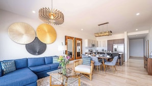 A modern living room with a blue sofa, a glass coffee table, and a dining area with wooden chairs. - Punta Serena | Los Tules Oceanview Penthouse Condo (Residencial Los Tules)