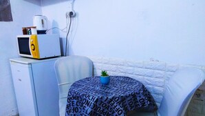 Una kitchenette pequeña con microondas, refrigerador y cafetera. Hay una mesa redonda con mantel estampado y una pequeña planta en maceta. La habitación tiene paredes blancas y piso de baldosas.