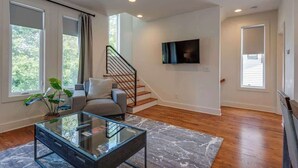 A modern living room with a glass coffee table, a grey sofa, a staircase, and a TV mounted on the wall.