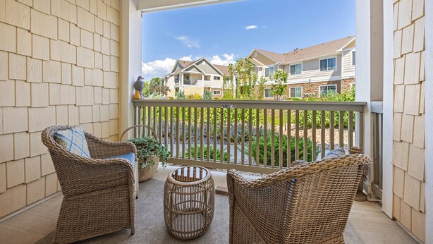 Een balkon met rieten meubels, een kleine ronde tafel en uitzicht op omliggende huizen.