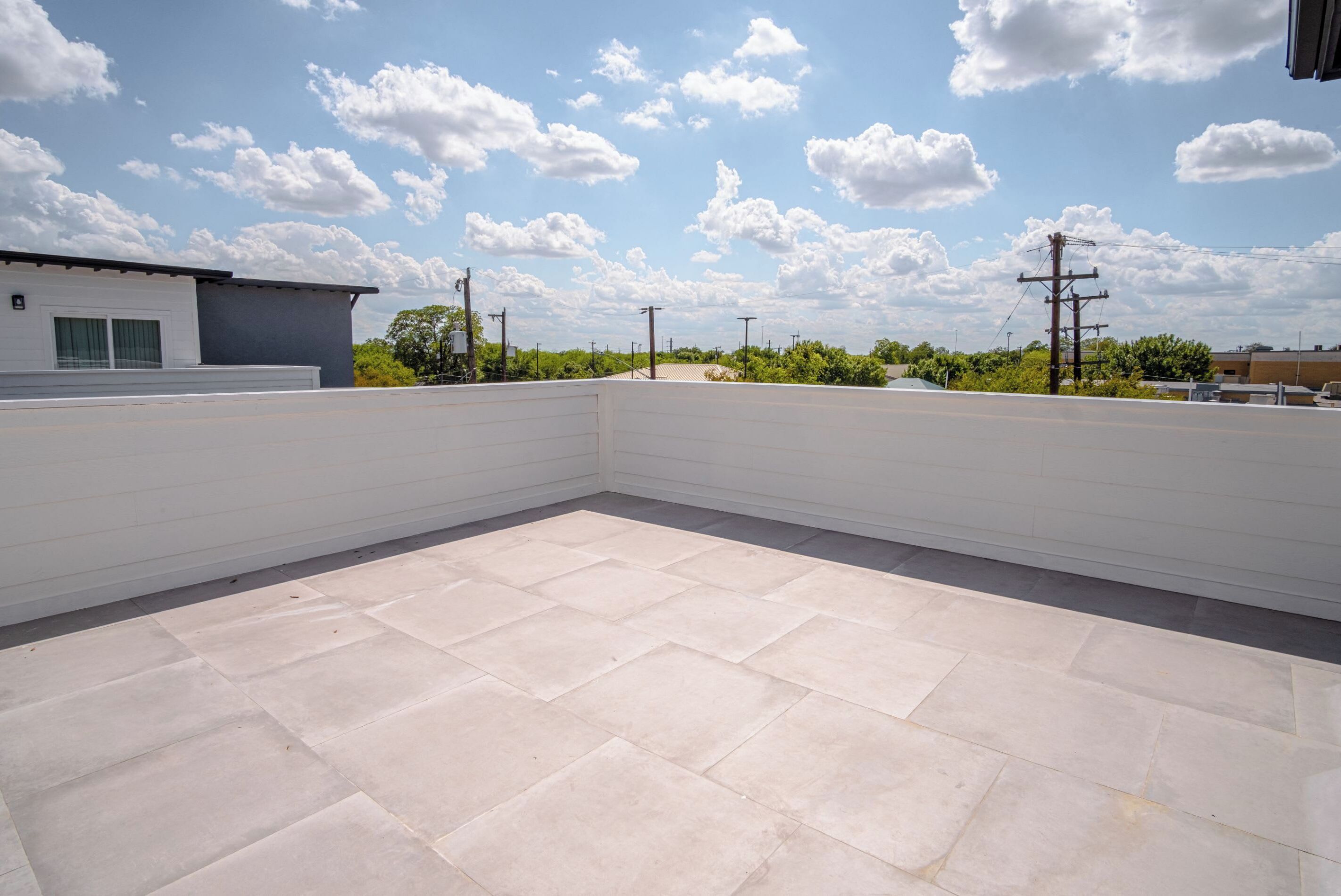 A rooftop terrace with a tiled floor, surrounded by a white wall and a clear sky with clouds.