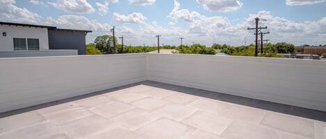 A rooftop terrace with a tiled floor, surrounded by a white wall and a clear sky with clouds.