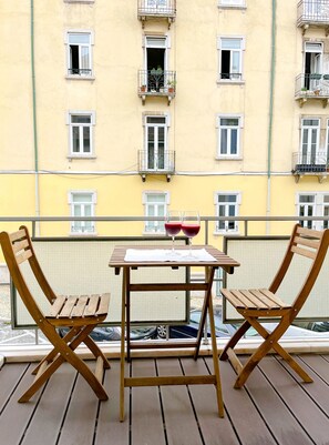 Ein Balkon mit zwei Holzstühlen und einem kleinen Tisch, mit Blick auf ein gelbes Gebäude mit zahlreichen Fenstern und Balkonen.
