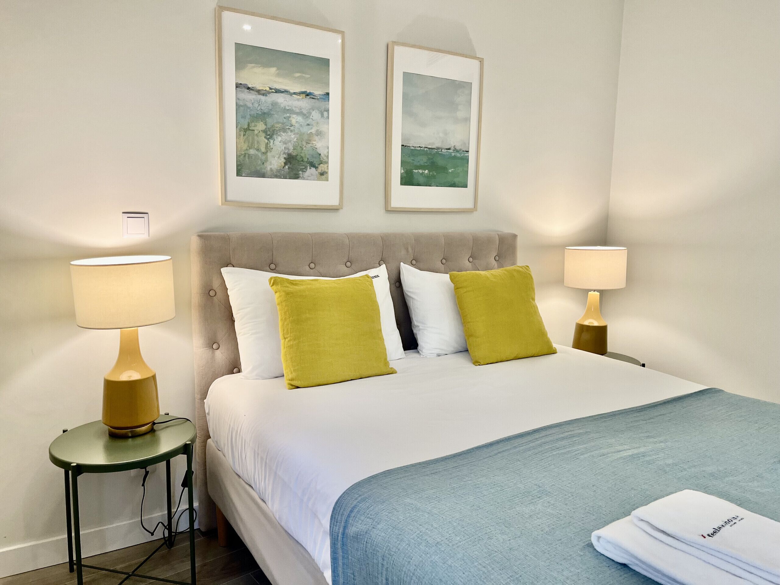A neatly made bed with white and green pillows, two framed landscape paintings above it, and two bedside lamps on matching nightstands.