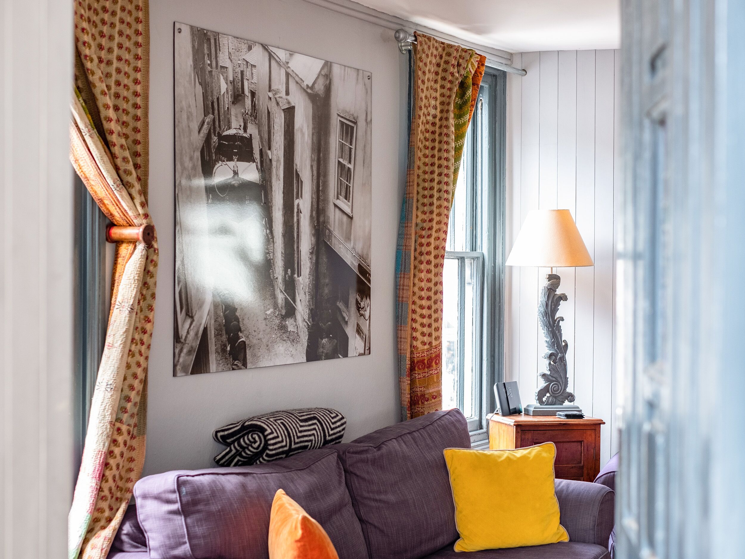 A living room with a purple sofa, a yellow pillow, a black and white photograph on the wall, and a lamp on a wooden side table.