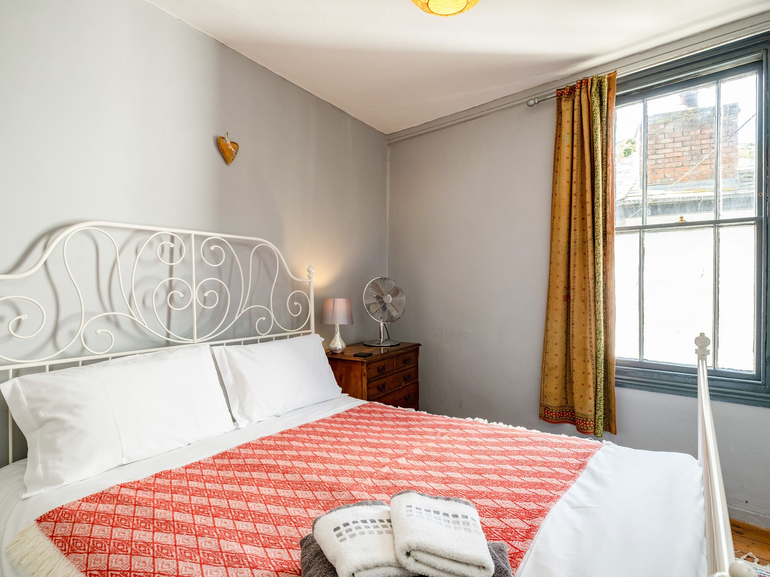 A neatly made bed with a red and white patterned bedspread, a white headboard, and a bedside table with a lamp and a fan.