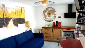 A modern living room with a blue sofa, a round wooden clock, and a kitchen area with a red table and various appliances.