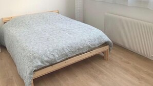 A single bed with a wooden frame and a grey knitted blanket in a room with a radiator and a window with blinds. - Agréable F2 Proche Gare (Le Mans)
