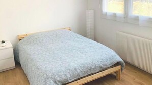 A bedroom with a wooden bed, a white nightstand, a radiator, and a window with a view of greenery outside. - Agréable F2 Proche Gare (Le Mans)