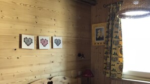 A wooden room with a bed, two heart-shaped wall art pieces, a nightstand, a lamp, and a window with patterned curtains.