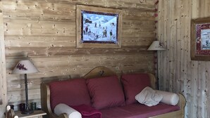 A cozy cabin interior with a wooden ceiling, a sofa with red and white checkered bedding, a wooden table with apples, and a framed picture on the wall.