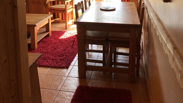A wooden dining area with a table and chairs, a red rug, and a view through a door to an outdoor balcony.