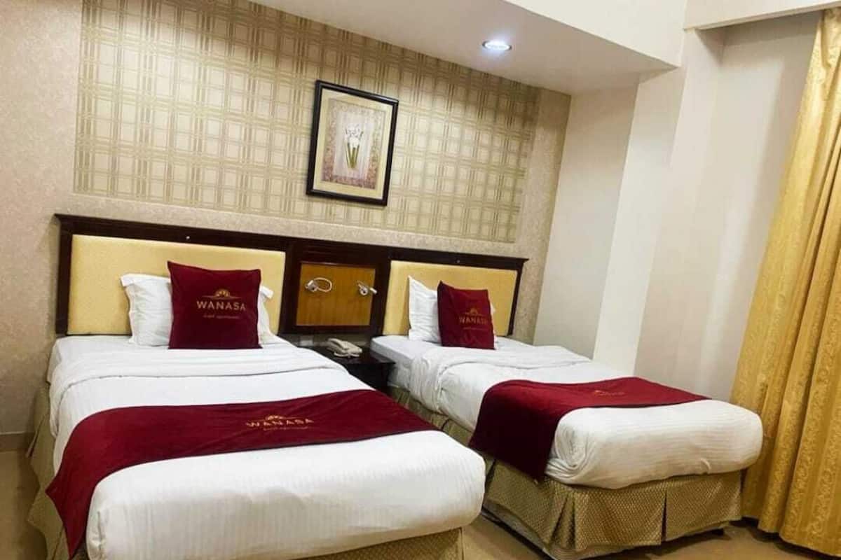 A hotel room with two beds, each with a red 'WANASA' pillow, a framed picture on the wall, and a gold curtain.