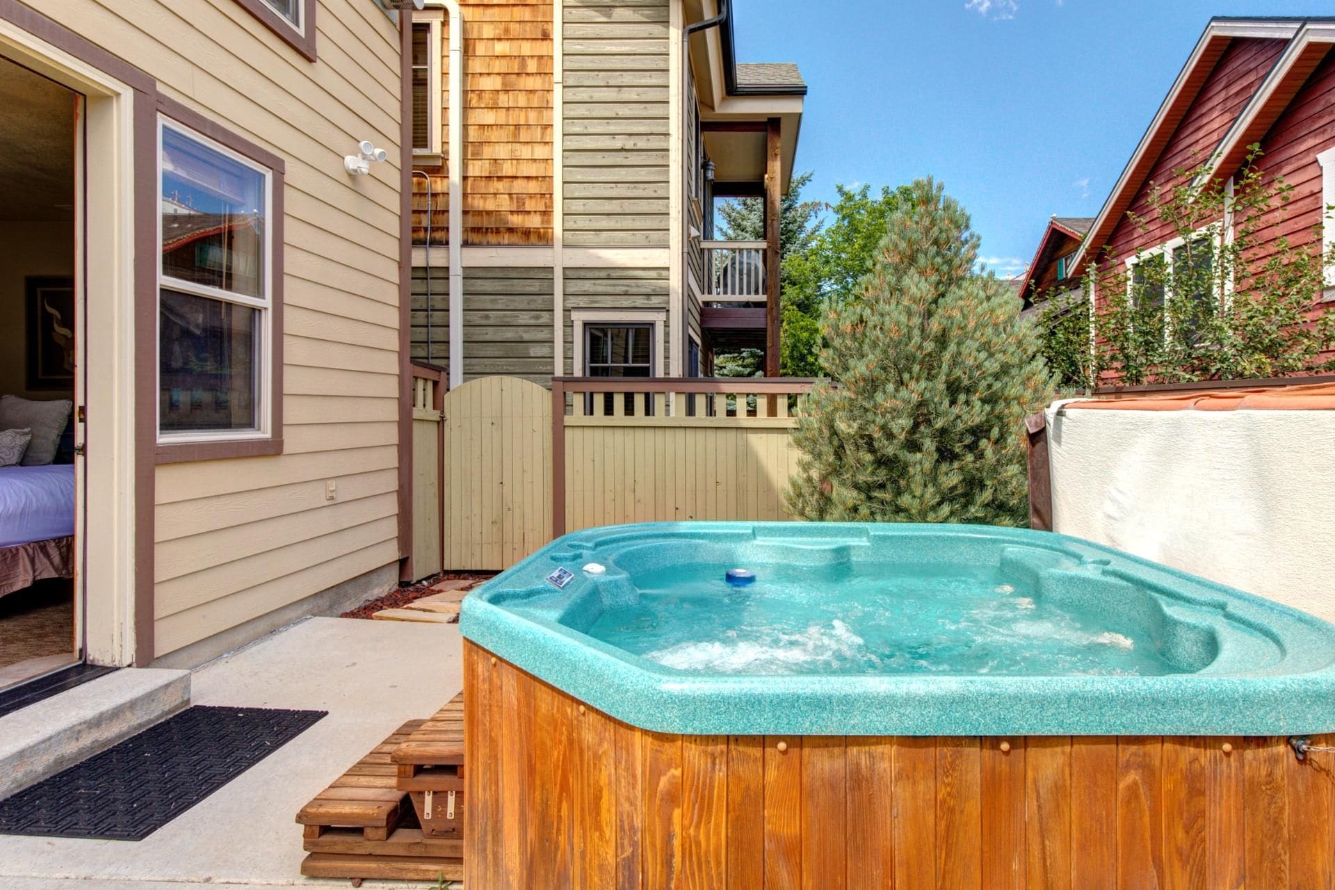 Un jacuzzi de madera en un patio trasero con cercado de madera, terraza de madera y una casa con revestimiento color beige y balcón.