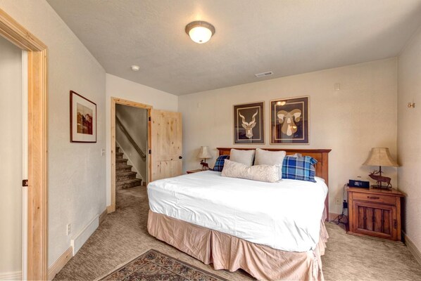 A bedroom with a large bed, a wooden headboard, two framed deer antlers on the wall, a nightstand with a lamp, and a staircase leading to another room.