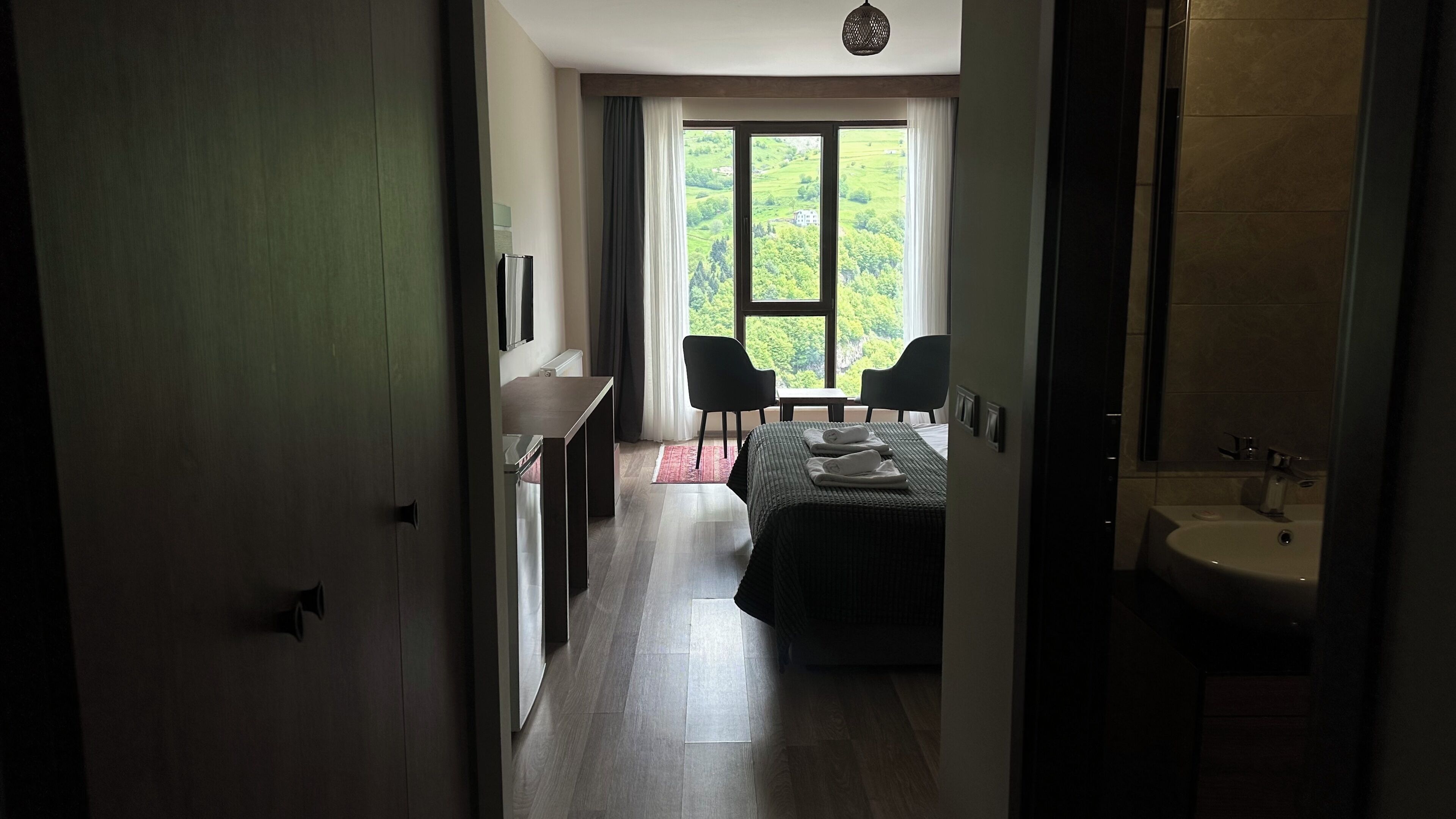 A hotel room with a bed, a desk, two chairs, a large window with a view of greenery, and a bathroom with a sink.