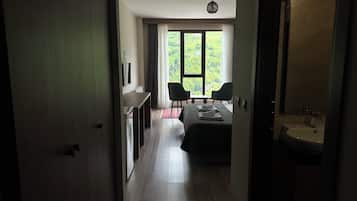 A hotel room with a bed, a desk, two chairs, a large window with a view of greenery, and a bathroom with a sink.