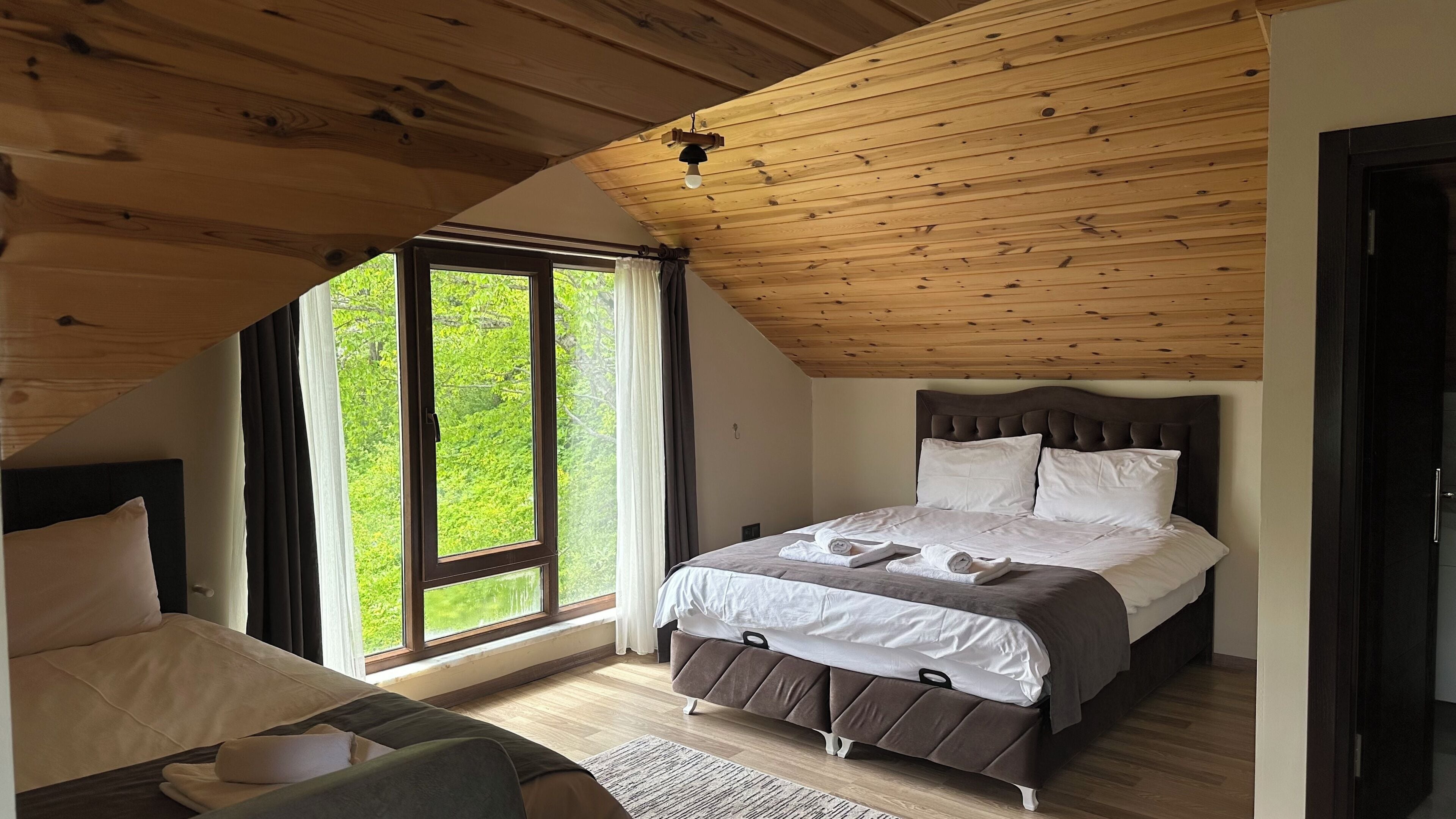 Een slaapkamer met schuine wand, een bed met hoofdbord, een raam met gordijnen en uitzicht op groen.