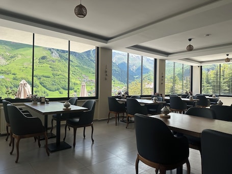 A dining area with tables and chairs, large windows with a mountain view, and ceiling lights.. Alya Teras Otel