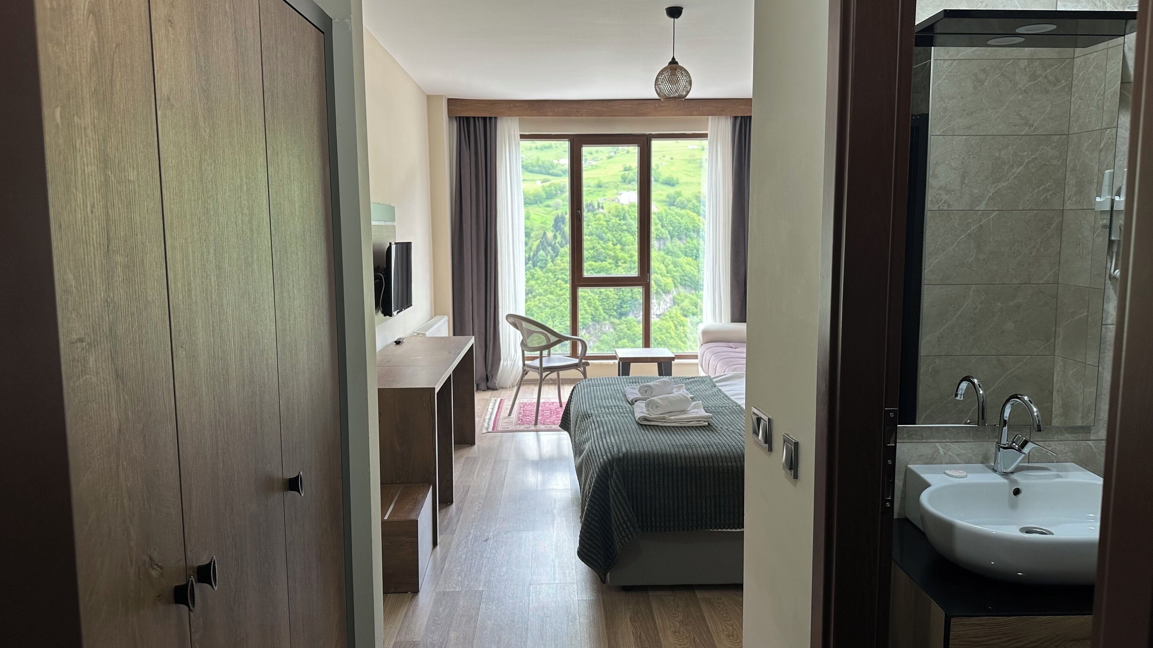 A bedroom with a bed, a desk, a chair, a window with a view of greenery, and a bathroom with a sink and mirror.