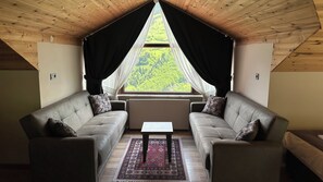 Ein gemütliches Dachzimmer mit hohen Decken, zwei Sofas, einem kleinen Tisch und Blick auf eine grüne Landschaft durch ein großes Fenster.