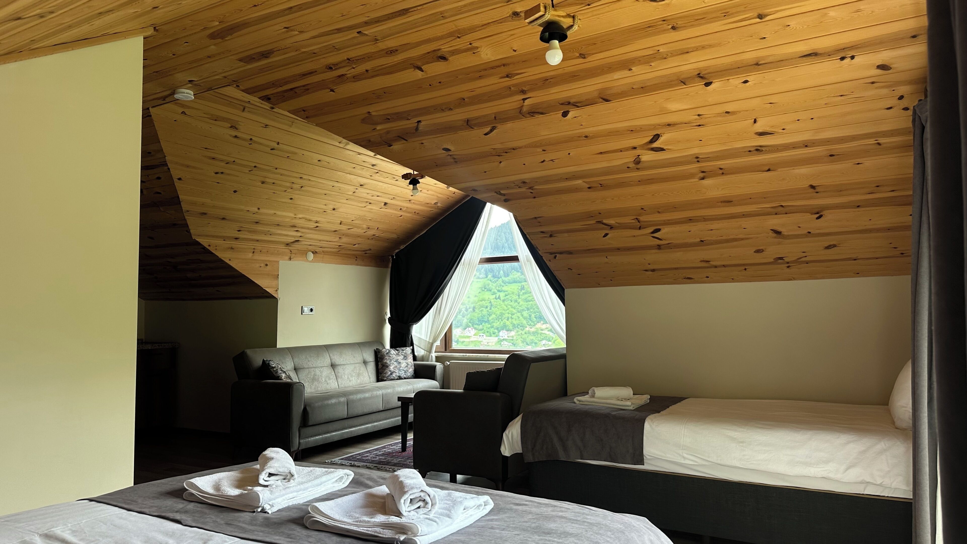Quarto com teto inclinado, sofá, cama, mesa com toalhas e janela com vista para o verde.