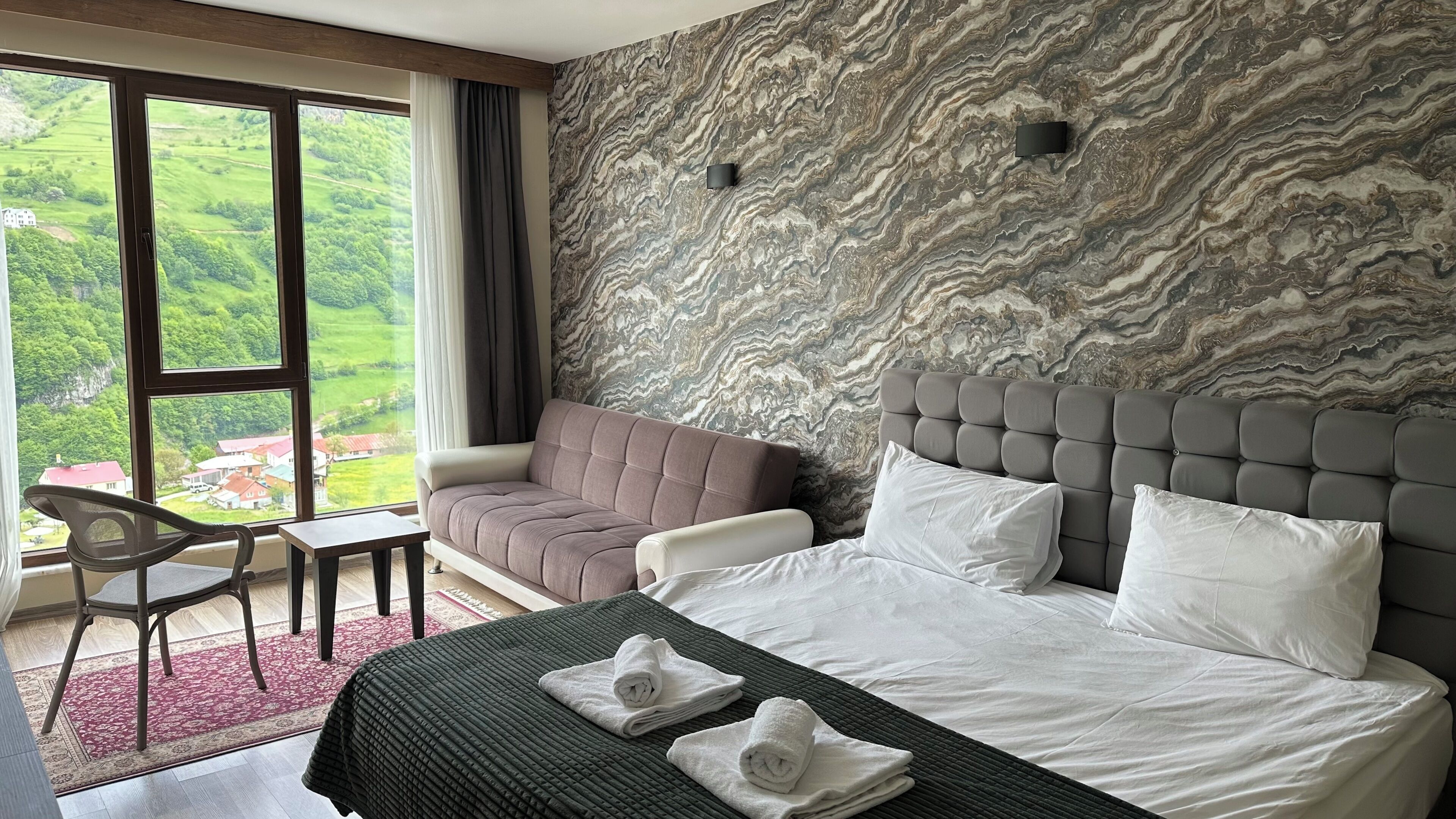 Habitación de hotel con cama, dos sillones, una mesita y vistas a zonas verdes a través de amplios ventanales, además de una pared con diseño de mármol.