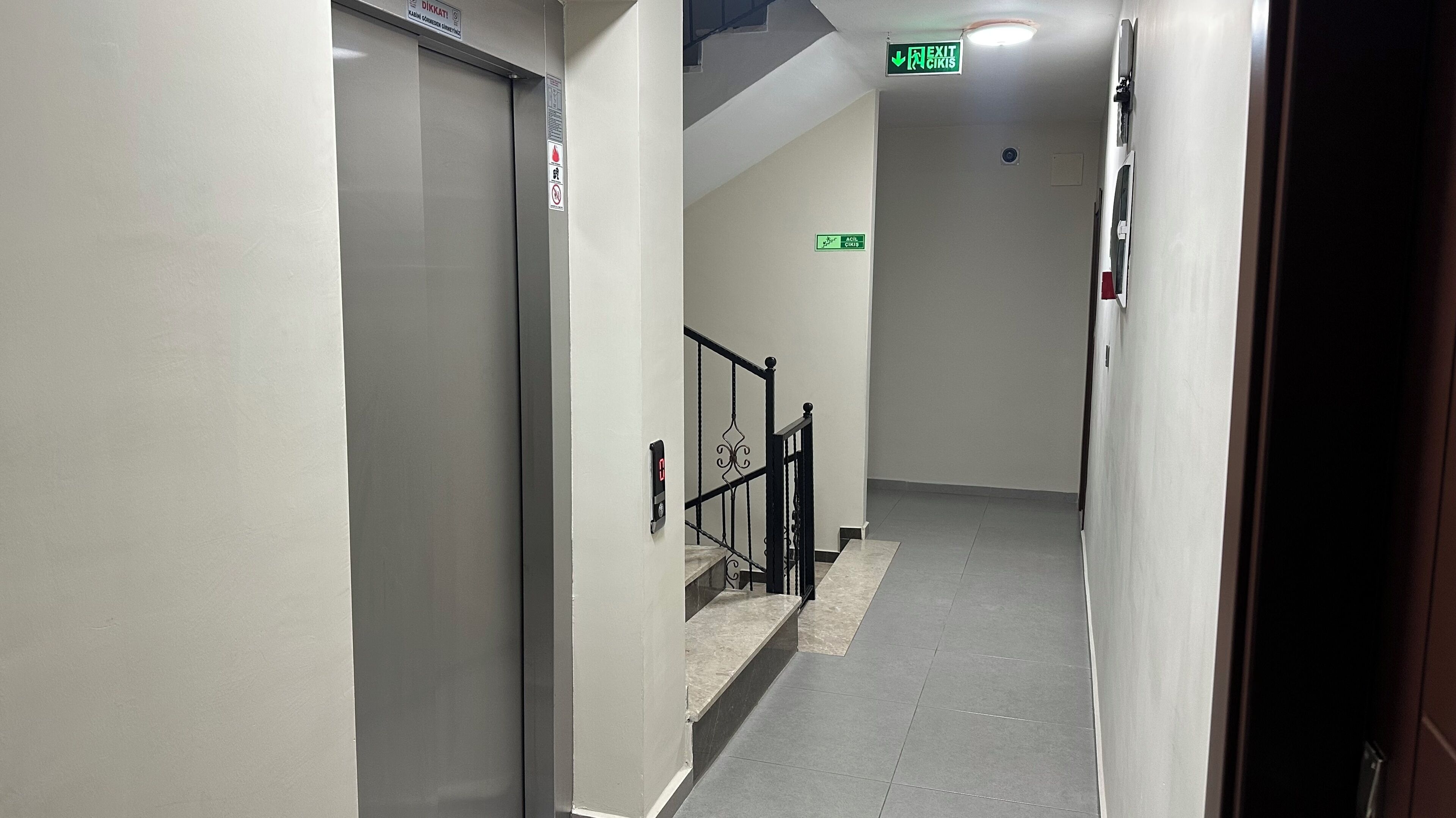 A narrow, well-lit hallway with a marble floor, a staircase with black railings, and an exit sign.