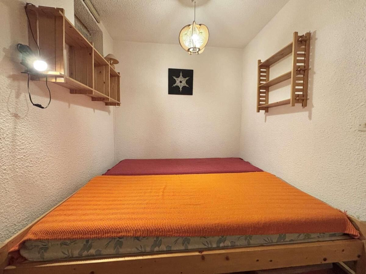 A small, single-bed room with a wooden bed frame, a red mattress, and a hanging light fixture.